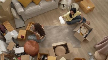 Oturma odasındaki mobilyaların taşınmasına yardım ederken yerde oturan mutlu oda arkadaşı. Yeni eve taşınmak için birlikte çalışan Asyalı lezbiyen aşığının havadan görünüşü. Yer değiştirme konsepti. Myrmidon.