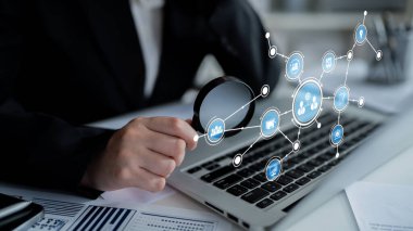A business professional uses a magnifying glass to analyze data displayed on a laptop in a modern workspace, showcasing concepts of digital connectivity and technology analysis. Tessel