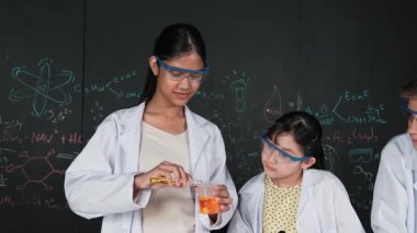 Değişik öğrenciler masada mikroskop ya da tüpü sıvıyla karıştırırken renkli solüsyonları karıştırıyorlar. Kimyasal teorisiyle karatahtanın önünde duran Asyalı kız deney tüpü tutuyor. Öğretim