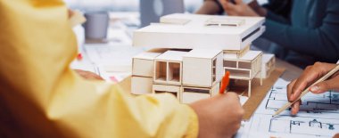 Bir grup profesyonel, modern bir çalışma alanında takım çalışması ve yaratıcılığı gösteren ölçekli bir model ve tasarım planları kullanarak bir mimari proje üzerinde işbirliği yapmaktadır. SACTR