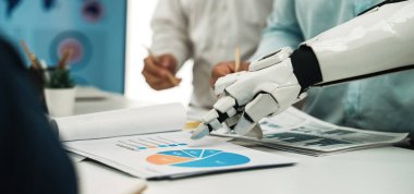 Robotik bir el, günümüz iş ortamında teknoloji ve yeniliklerin kesişiminin gösterildiği modern ofis ortamındaki iş haritalarına işaret eder. Noogenesis