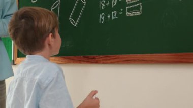 Düşük açılı kamera, yetenekli öğretmen çocuğa bakarken beyaz çocuğun tahtaya cevap yazdığını gösteriyor. Sorulara cevap vermek için gönüllü olan zeki ilkokul öğrencilerini kapatın. Eğitim. Pedagoji.