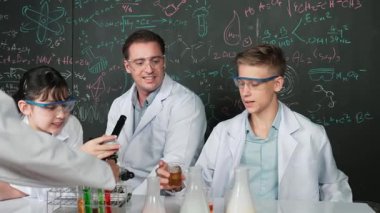 Laboratuvar önlüğü giyen genç bir çocuk, STEM fen sınıfında tahtada renkli sıvıyla bekletiliyor. Öğretmen masada tavsiye verirken bir grup farklı öğrenci deney yapıyor. Öğretim