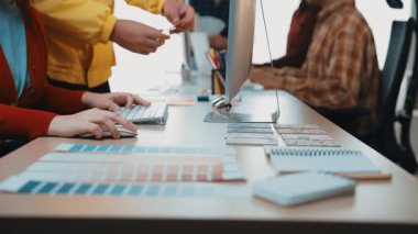 A diverse team collaborates on a computer project in a creative office environment. Various color swatches and notebooks detail their innovative brainstorming efforts. SACTR