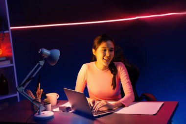 Geceleri sosyal medyada canlı yayında pozitif podcastla konuşan gülümseyen genç, güzel Asyalı etkileyici pembe neon ışıklı modern stüdyoda sunum için dizüstü bilgisayar kullanıyor. Taktik.