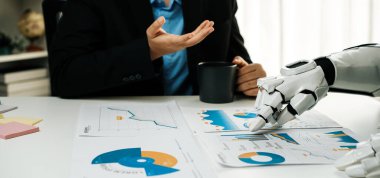 Bir insan ve bir robot ofis masasında etkileşime girer, kahve içerken çizelgeleri ve veri raporlarını birlikte inceler, iş analizlerinde modern işbirliği sergilerler.