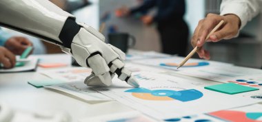 Bir robot el, modern bir ofiste veri grafiklerini ve grafiklerini analiz etmek için insan eliyle işbirliği yapar. İş dünyasındaki teknoloji ve takım çalışmasının karışımını açıklar. Noogenesis