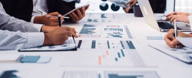 Bir iş toplantısında yer alan çeşitli profesyonellerden oluşan bir grup, takım çalışmasını vurgulayarak, modern bir ofis ortamında kağıt ve cihazlar üzerinde sunulan tablo ve grafikleri analiz ediyor. SACTR