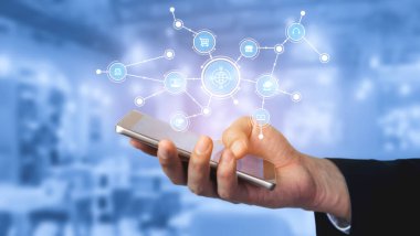 Akıllı telefonu tutan bir el, modern iletişim ve teknoloji entegrasyonunu sembolize eden dijital ağ simgelerini gösteren mavi bir arkaplan karşısında sergilenir. Tessel