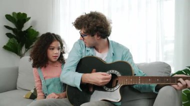 Beyaz bir baba, tatlı kızına koltukta otururken gitar çalmayı öğretiyor. Müzik öğrenirken babasına gitar çalarken bakan tatlı öğrenci kız. Aile eğlence konsepti. Pedagoji.