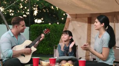Baba gitar çalarken anne ve kız birlikte kampta oturuyorlar. Ebeveynler, genç nesil çapraz kuşak farklılıklarıyla zaman geçirmek için açık hava kamp aktivitelerini kullanıyorlar. Uyumsuzluk.