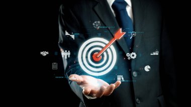 Kendine güvenen bir işadamı dijital bir hedefe kırmızı bir okun merkeze isabet ettiğini gösterir, başarıyı simgeler ve siyah bir arkaplana karşı kurumsal stratejiye odaklanır. Dörtgen