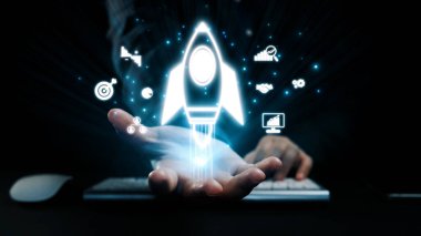 Bir el, yenilik ve büyümeyi simgeleyen, karanlık bir ofis zeminine kurulmuş, teknoloji ve iş gelişiminin yolculuğunu gösteren parlayan bir roket tutuyor. Dörtgen