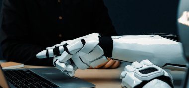 Günümüz dijital dünyasında insanlar ve gelişmiş teknolojiler arasındaki işbirliğini gösteren, modern çalışma alanındaki bir dizüstü bilgisayarı gösteren bir robot elin yakın çekimi. Noogenesis