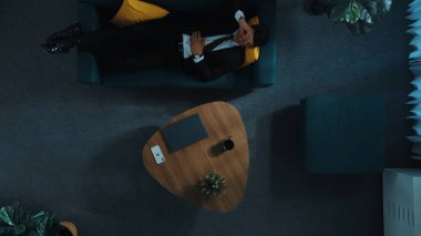 Stresli yöneticinin telefon kullanarak takımla konuştuğu yukarıdan aşağıya hava görüntüsü. İş adamı iş arkadaşlarını arıyor. Dizüstü bilgisayarda çalışırken ve kanepede yatarken. Uzaktan kumanda. Zaman aşımı. Müdürlük.