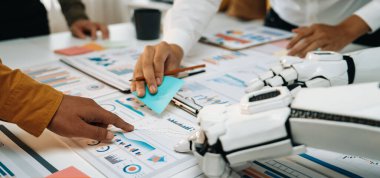 Bir insan ve bir robot, renkli yapışkan notlar ve detaylı belgelerle çevrili, modern ofis ortamında takım çalışması ve yenilik içeren bir proje üzerinde işbirliği yapıyorlar.