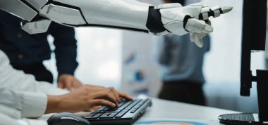 İnsanlarla makineler arasındaki işbirliğini vurgulayarak, modern bir ofiste bir kişiye klavyede yardım eden robotik bir kolun yakın çekimi. Noogenesis