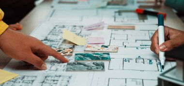 Ekip planlama oturumunda el ele tutuşup çizimler ve görsel yardımlarla mimari bir düzene işaret ediliyor. Katılımcılar proje fikirleri üzerinde aktif olarak işbirliği yapıyorlar. SACTR