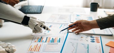 Bir insan ve robot, modern bir ofis ortamında finansal tablolar ve raporlarla veri analizi yaparak iş birliği yapıyor ve yenilikler sergiliyor.