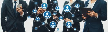 Resmi giyinmiş bir grup profesyonel, dijital cihazlar aracılığıyla iş ağı kuruyor ve iletişimi sergiliyor. Simgeler bağlantıları ve işbirliğini temsil ediyor. Dostluk