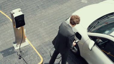 İlerici işadamı, şehir merkezinde dolaşmadan önce şarj cihazından elektrikli arabasına şarj prizi takıyor. Çevre dostu şarj edilebilir araba, sürdürülebilir ve temiz enerjiyle çalışıyor..
