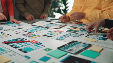 A diverse team of designers engages in collaborative discussion, using colorful sticky notes and mobile mockups to strategize user experience in a modern workspace. SACTR
