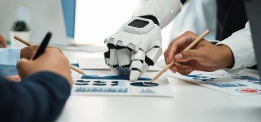 Bir insan eli ve bir robot el, günümüz dijital dünyasında takım çalışması ve teknolojik yeniliklerin altını çizerek, modern ofis alanındaki veri raporlarını işbirliği içinde analiz eder.