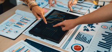 Modern bir ofiste çeşitli iş haritaları ve raporlarla çevriliyken, dijital tablet etrafında çalışan dinamik bir profesyonel grup veri analizine katılıyor. SACTR