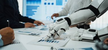 Bir robot el, bir iş toplantısı sırasında çizelgeleri ve grafikleri analiz eder. Profesyoneller, veri odaklı içgörüler ve gelecekteki stratejileri tartışmak için masa etrafında işbirliği yaparlar.